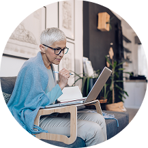 Person with short gray hair working on a laptop at home with notebook and pen.
